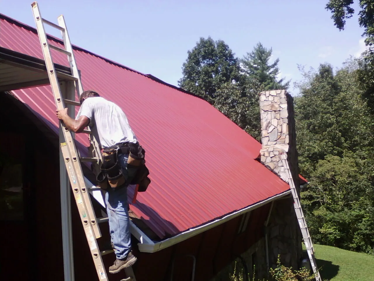 Roofer on a ladder beside a red metal roof and stone chimney — Soffit & Fascia Repair by Roofing Brothers in Skull Valley